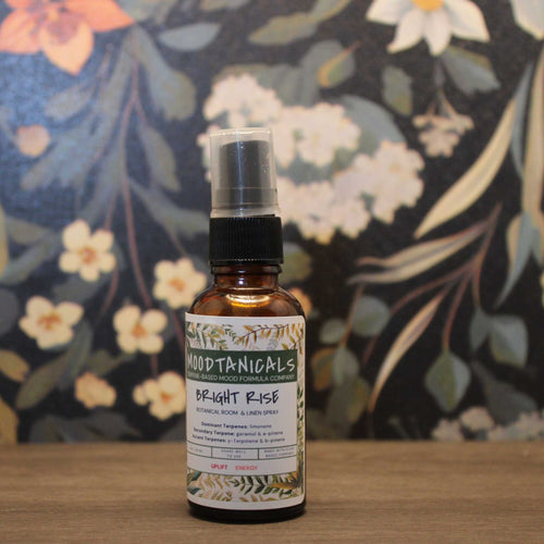 Bottle of Moodtanicals 'Bright Rise' room spray on a wooden surface with a floral background.