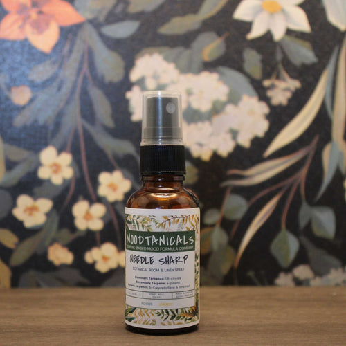 Bottle of Moodtanics Needle Sharp spray on a wooden surface with a floral background