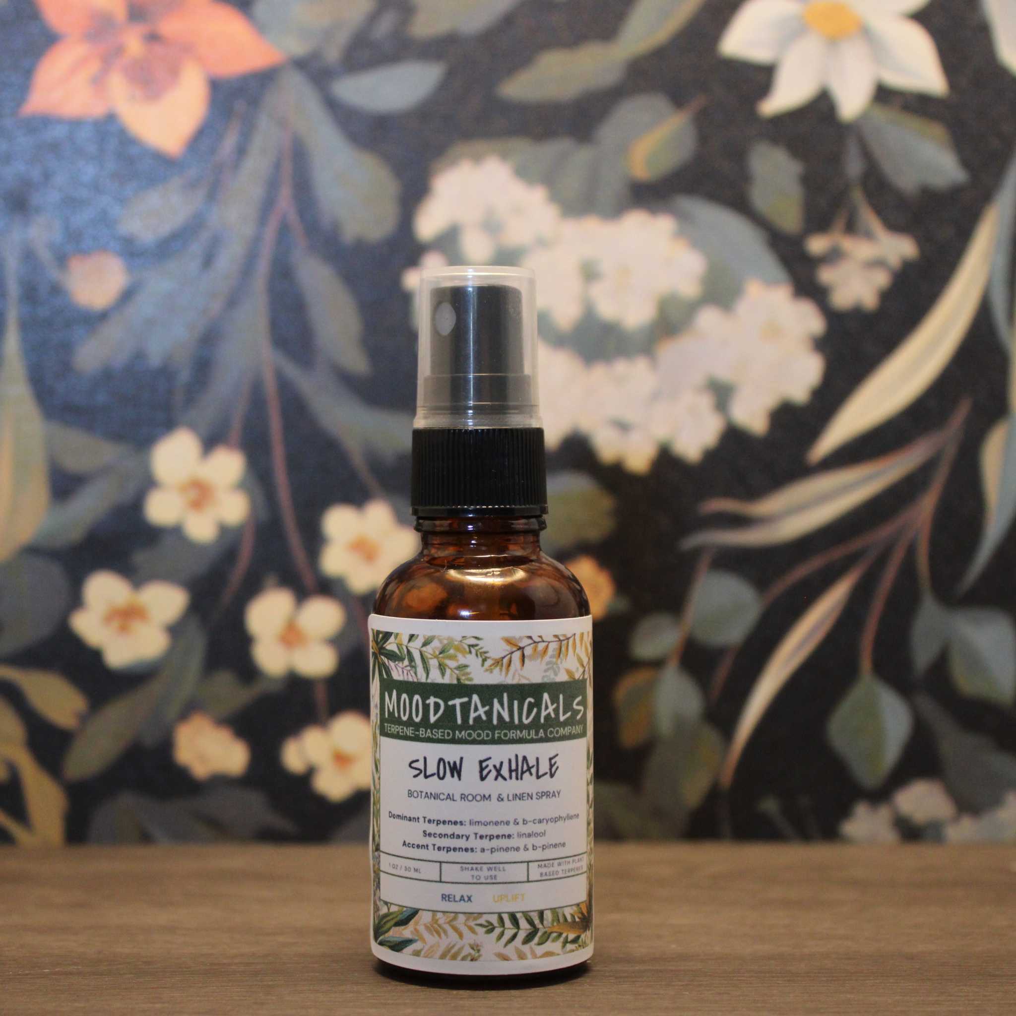 Bottle of Moodtanicals Slow Exhale product on a wooden surface with a floral background